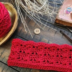Red crochet earwarmer displayed on a wooden surface with matching yarn, crochet hook, stitch markers, and rustic fall accents including dried wheat and pinecones.