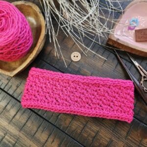 Flat lay of a pink crocheted earwarmer with textured stitches, surrounded by yarn, scissors, stitch markers, and a handmade crochet tag.