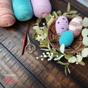 Styled flat lay on a dark wood surface featuring three finished crocheted amigurumi eggs displayed in a woven basket surrounded by eucalyptus stems and white berry sprigs. The eggs show three variations — lavender with purple French knot speckles, blush pink with cream stripes worked in seed stitch, and solid teal. In the upper left three yarn cakes in blush pink, mint green, and soft lavender are arranged alongside a dark wooden crochet hook and decorative scissors. Speckled Nest Eggs Set — Week 3 of the Stitch the Season: Spring Awakening CAL by Mistie Bush of American Crochet.
