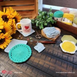 Styled flat lay on a dark wood surface showing the complete Spring Coaster Set collection in progress. Four coaster designs are displayed — a large mint green round coaster with lace border detail, a gray square coaster, a small gray square coaster in progress, and two flower and hexagon coasters in yellow and sage green with white borders. An American Crochet branded white mug sits on a finished square coaster in the upper left alongside bright yellow sunflowers. A wooden heart shaped yarn bowl holds pale blue cotton yarn with a dark wooden crochet hook attached to a coaster in progress. Colorful stitch markers and decorative copper scissors are scattered nearby. A dark wood tray in the upper right holds yarn cakes in mint green, blush pink, sage green, yellow, and white alongside lush eucalyptus. Spring Coaster Set — Week 4 of the Stitch the Season: Spring Awakening CAL by Mistie Bush of American Crochet.